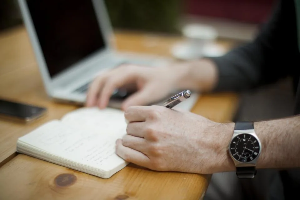 Manos tomando notas en un cuaderno junto a una computadora portátil